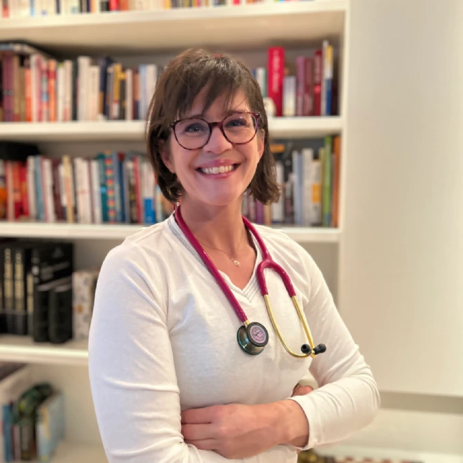 Dr. med. Anna Kocsis mit Stethoskop, lächelnd vor einer Bücherwand.