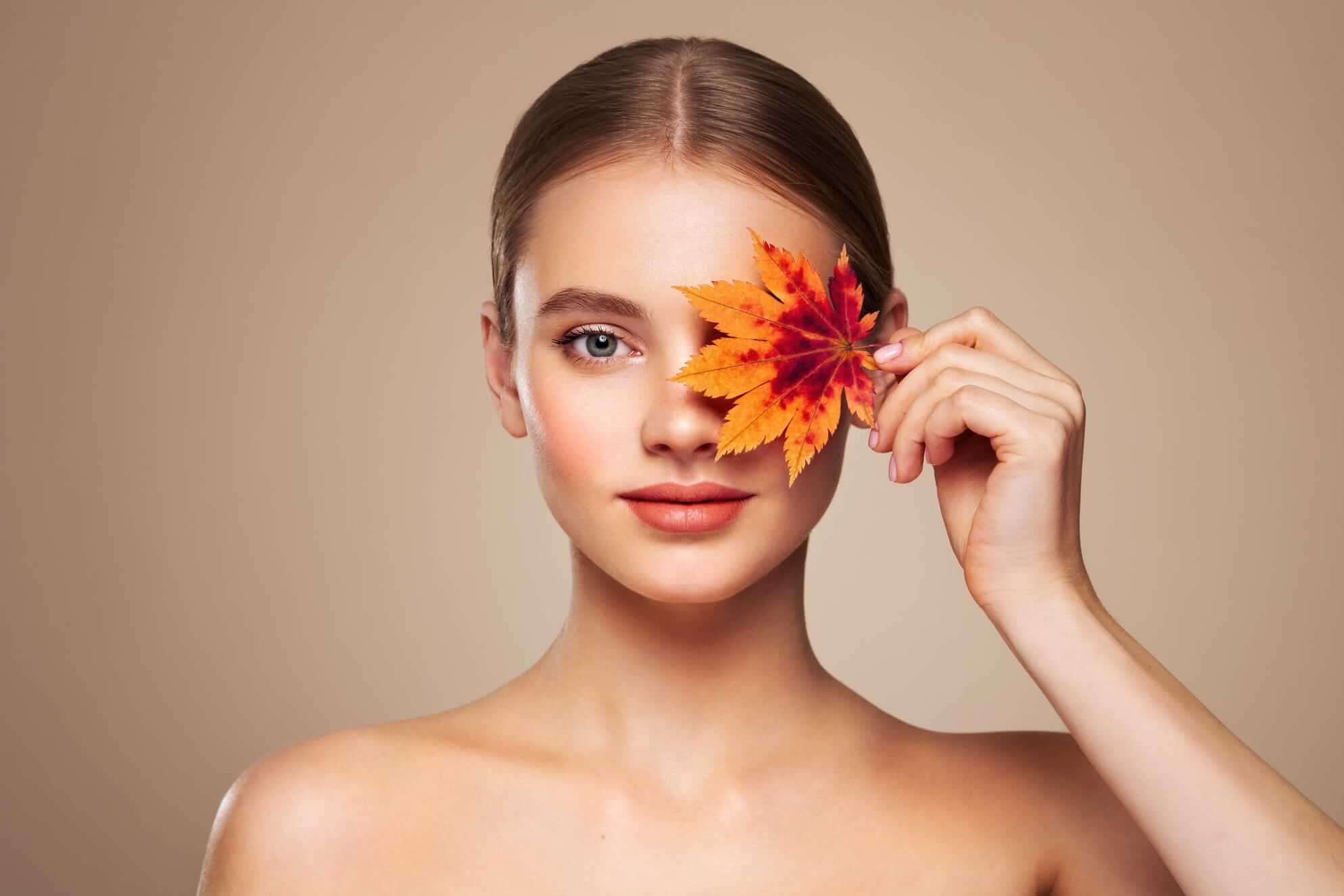 Junge Frau mit einem bunten Herbstblatt vor einem Auge, lächelnd vor einem beige-braunen Hintergrund.
